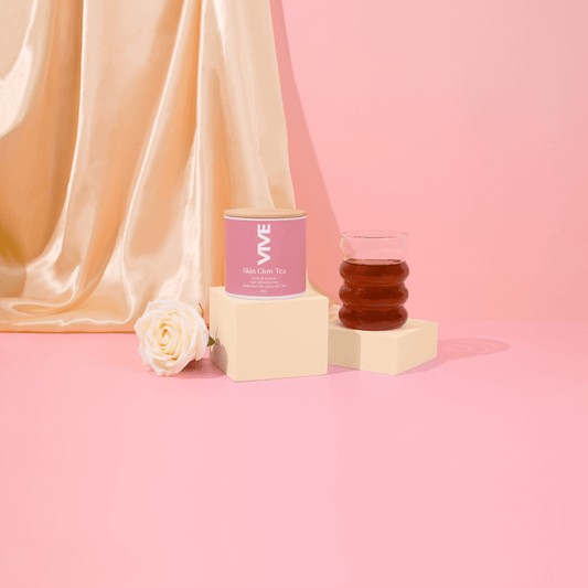Skin Glow Tea canister next to a glass of tea and a flower on a pink background, showcasing an infusion for glowing skin.